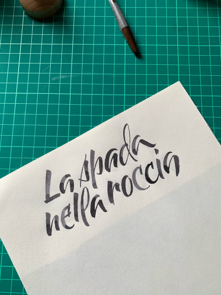 A piece of white paper on a green grid mat. A calligraphy brush can be seen on the upper border. Calligraphy text on paper: "la spada nella roccia" or "the sword in the stone."